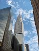 Ground-level view of an 80-story building; the structure has a stone, whitish exterior with several setbacks. Statues project from the building near the 60th floor, and the building tapers into a thin spire containing angled triangular windows.